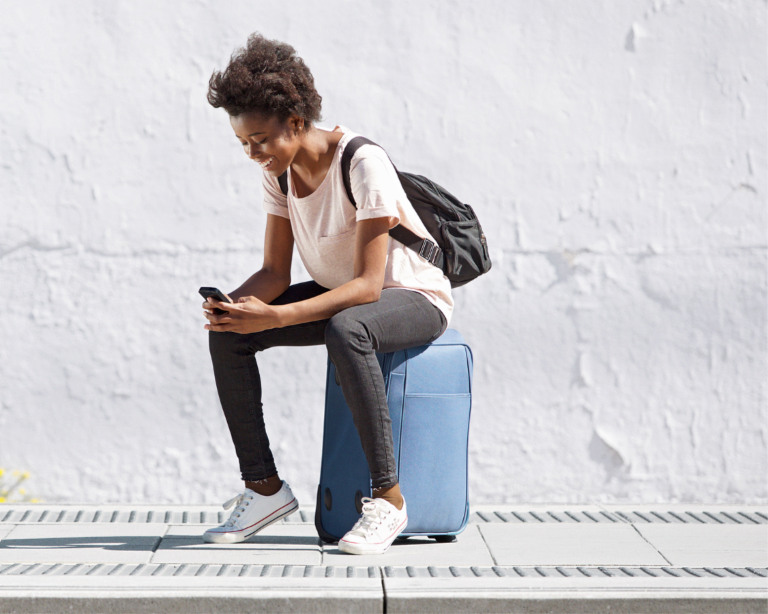 Eine junge Frau sitzt auf einem Koffer, schaut auf ihr Mobiltelefon und lächelt.